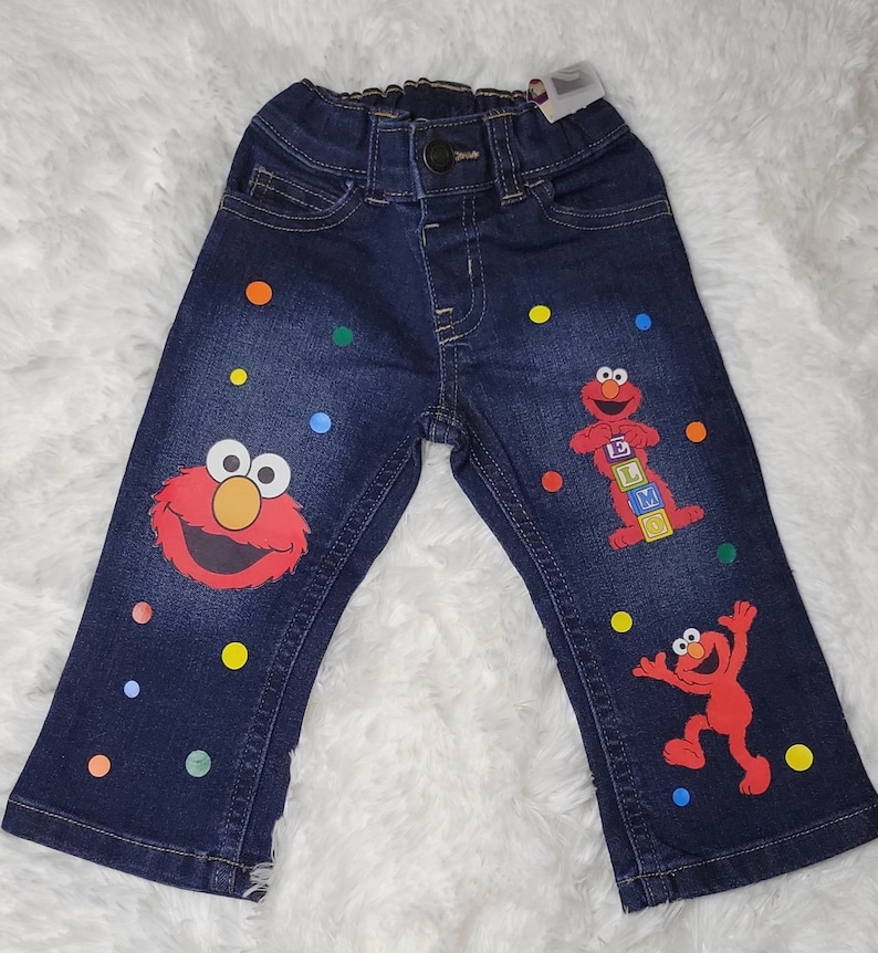 May include: Blue denim jeans with colorful polka dots and a red Sesame Street character, Elmo, printed on the legs. Elmo is holding blocks that spell "ELMO".