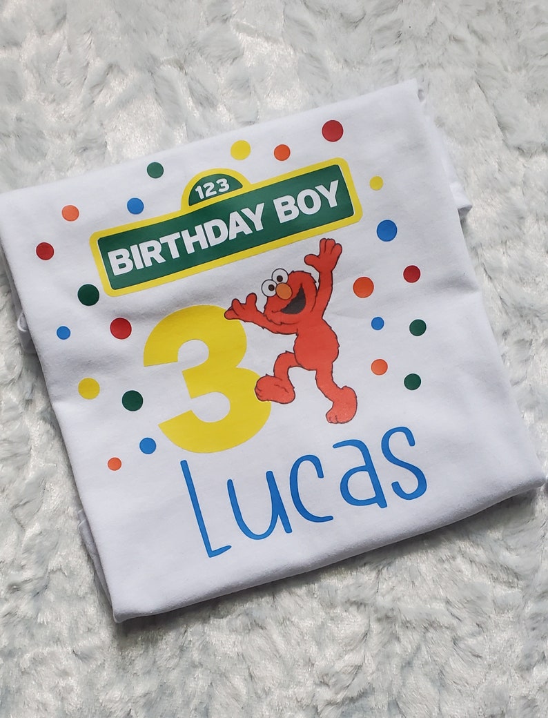 May include: White t-shirt with a Sesame Street theme. It features a red Elmo character, a yellow number 3, and a green sign that says "Birthday Boy" with the number 123. The name Lucas is written in blue below the number.