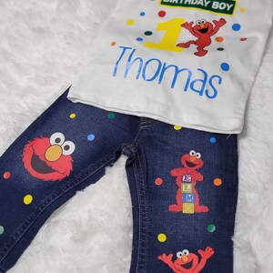 May include: A white t-shirt with a red Sesame Street character, a yellow number one, and the text "Birthday Boy" on a green sign. The shirt also has the name "Thomas" in blue text. The t-shirt is paired with blue jeans with a red Sesame Street character and colorful dots.