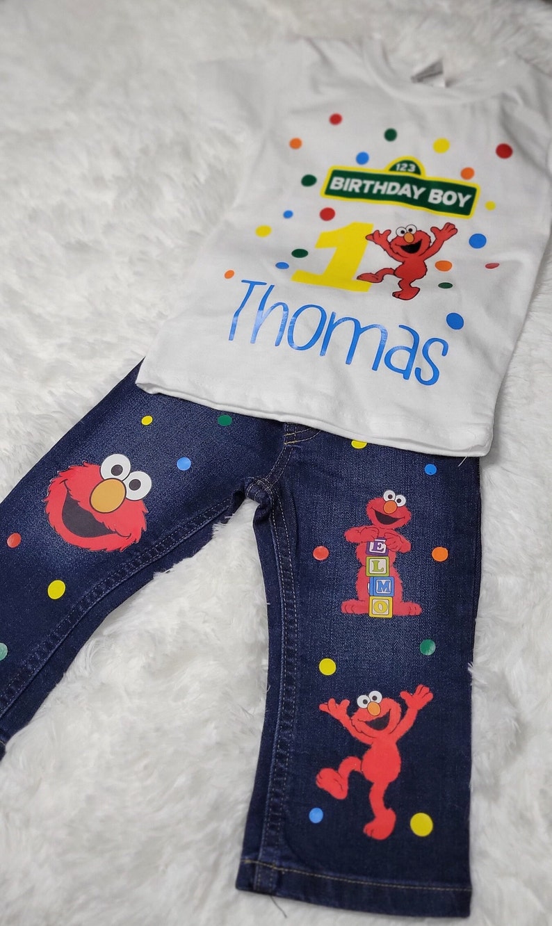 May include: A white t-shirt with a red Sesame Street character, a yellow number one, and the text "Birthday Boy" on a green sign. The shirt also has the name "Thomas" in blue text. The t-shirt is paired with blue jeans with a red Sesame Street character and colorful dots.