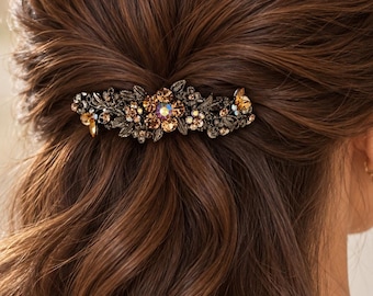 May include: A decorative hair clip featuring an intricate dark metal floral design adorned with amber and iridescent crystals. The accessory is secured in long, wavy brown hair, showcasing a detailed arrangement of leaves and sparkling stones.