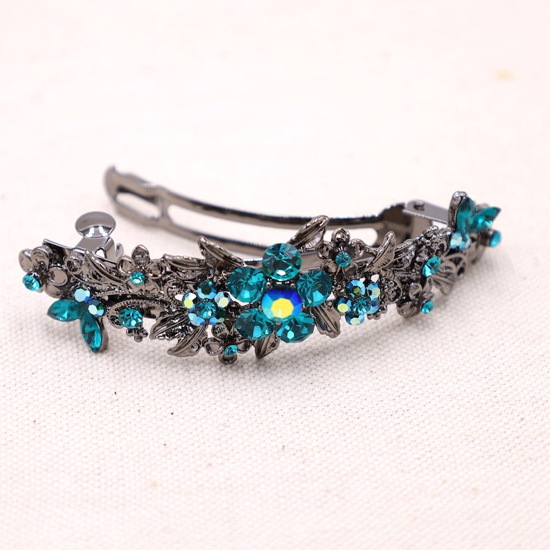 May include: A dark gunmetal hair clip featuring an intricate floral design adorned with multiple teal-colored rhinestones. The central flower is highlighted by a larger, iridescent teal crystal that reflects light, set against a metallic filigree base.