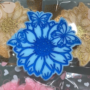 May include: A blue and light blue flower-shaped decoration with butterfly accents. The flower has detailed petals and is surrounded by other decorative items in clear packaging. The overall theme is floral and decorative.