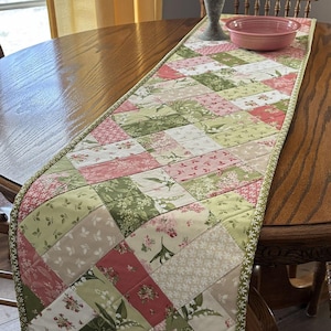 May include: A quilted table runner with a patchwork design. The runner features squares of floral fabric in shades of pink, green, and cream. A green border with a scalloped edge frames the runner, which is placed on a wooden table.