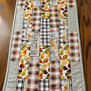 Puede incluir: Camino de mesa acolchado rectangular con diseño de patchwork. El camino de mesa presenta franjas verticales alternas de cuadros, estampados de hojas y gráficos de camiones. Los colores incluyen naranja, marrón, gris y blanco. Los bordes están acabados con un estampado floral gris.