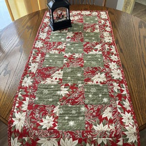 May include: A festive Christmas table runner with a red and green poinsettia design. The runner is on a round wooden table, with a black lantern containing a Santa figurine placed on top.