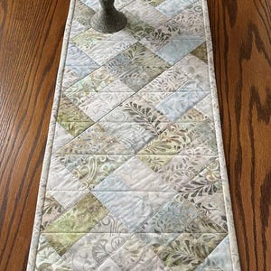 May include: A rectangular quilted table runner featuring a geometric pattern of diamond-shaped fabric patches in soft shades of sage green, pale blue, and white, decorated with various botanical leaf and swirling vine prints.