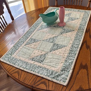 May include: A rectangular quilted table runner with a floral and geometric pattern. The runner features a light green and beige colour scheme with a border of green floral fabric. A turquoise jug and pink vase sit on top.