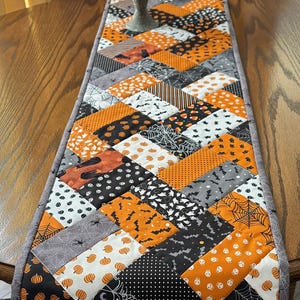May include: A rectangular table runner featuring a patchwork design with a herringbone pattern. The fabric squares display various Halloween-themed prints including orange pumpkins, black spiderwebs, bats, candy corn, and white skulls on black, orange, and white backgrounds, finished with a gray border.