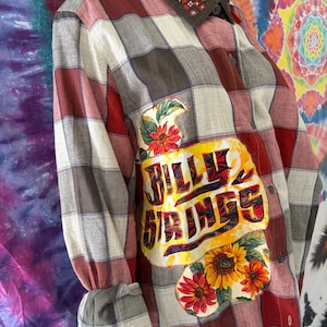 Könnte beinhalten: Ein langärmliges Button-Down-Hemd mit einem Karomuster in Rot-, Grau- und Weißtönen. Das Hemd hat einen dekorativen Aufnäher mit dem Text "BILLY SPRINGS" und Sonnenblumenakzenten. Der Kragen ist braun mit floraler Stickerei.