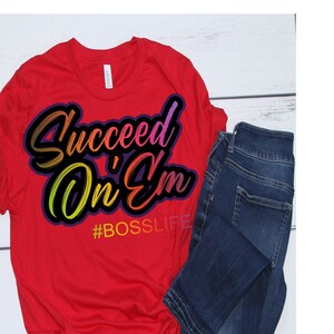 Può includere: Maglietta rossa con una grafica colorata che dice "Succeed On 'Em" e l'hashtag "#BOSSLIFE".