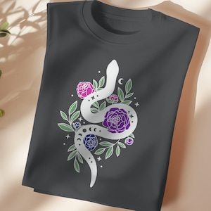 May include: A dark gray t-shirt with a white snake design. The snake is curled around a cluster of purple and pink flowers. The design is surrounded by small white stars and crescent moons.