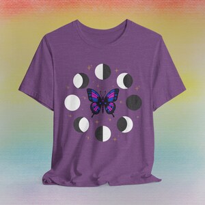 May include: A purple t-shirt featuring a butterfly with blue, pink, and purple wings, surrounded by phases of the moon. The design includes gold stars. The shirt is made of a soft material.