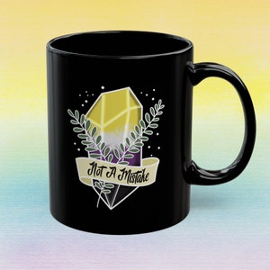 May include: Black ceramic mug featuring a yellow, purple, and white crystal design with green leaves and the text "Not A Mistake" on a banner. The mug has a curved handle.