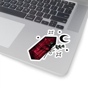 Könnte beinhalten: Ein roter Kristall mit einem grünen, blättrigen Stiel und einem Halbmond, umgeben von weißen Sternen auf einem silbernen Laptop.