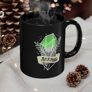 Puede incluir: Taza de cerámica negra con un diseño de cristal verde y blanco y el texto "Not A Mistake" en una pancarta amarilla.
