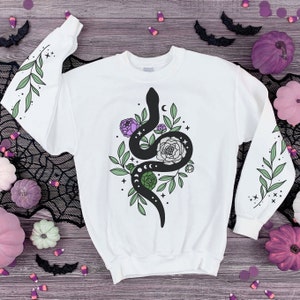 Puede incluir: Sudadera blanca con un diseño de serpiente negra, rodeada de hojas verdes y flores moradas. La serpiente está enroscada alrededor de una luna y estrellas.
