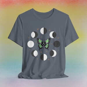 May include: A slate blue t-shirt featuring a butterfly with green and black wings, surrounded by a circle of moon phases. The design includes white, black, and gray moons, with small star accents.