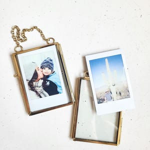 May include: Two gold-colored metal photo frames with attached chains, holding instant photos. One frame displays a photo of a person kissing a child, the other shows a desert landscape with a saguaro cactus. The third frame is empty.