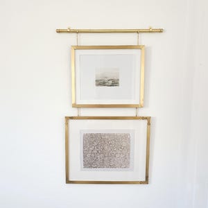 May include: Two framed prints hanging from a gold-colored rod. The top frame contains a grayscale image of a seascape. The bottom frame displays a detailed floral pattern. Both frames are rectangular with gold borders and are suspended by chains.