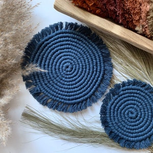 May include: Two navy blue macrame coasters with a spiral design and fringe detail. The coasters are round and sit on a white surface with dried grasses and a wooden tray in the background.