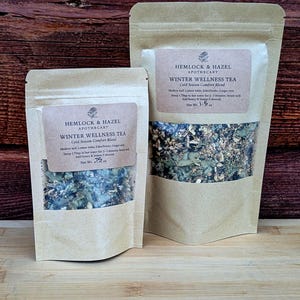 May include: Two kraft paper pouches of Hemlock & Hazel Apothecary Winter Wellness Tea. The smaller pouch contains 0.75 oz of tea, and the larger pouch contains 1.5 oz. Each pouch has a label with the tea's name and ingredients.