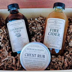 May include: A gift set featuring three apothecary products: Elderberry Syrup, Fire Cider, and Chest Rub. The bottles and tin are labeled with the brand name "Hemlock & Hazel". The Fire Cider bottle contains 8 oz (237 mL). The Chest Rub tin contains 2 oz (56g).