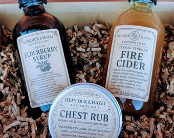 May include: A gift set featuring three apothecary products: Elderberry Syrup, Fire Cider, and Chest Rub. The bottles and tin are labeled with the brand name "Hemlock & Hazel". The Fire Cider bottle contains 8 oz (237 mL). The Chest Rub tin contains 2 oz (56g).