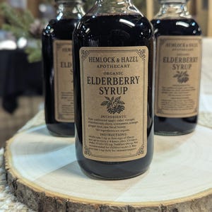 Pu&ograve; includere: Tre bottiglie di vetro di sciroppo di sambuco biologico Hemlock & Hazel Apothecary. Ogni bottiglia ha un tappo nero e un'etichetta marrone con testo. Le bottiglie sono riempite con un liquido viola scuro e sono esposte su una fetta di legno.