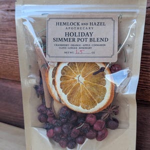 May include: A clear, sealed bag containing a holiday simmer pot blend. The bag includes dried orange slices, cranberries, cinnamon sticks, and star anise. The label reads "Hemlock and Hazel Apothecary Holiday Simmer Pot Blend". Net weight is 1.5 oz.