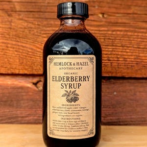 Pu&ograve; includere: Una bottiglia di vetro scuro di sciroppo di sambuco biologico Hemlock & Hazel Apothecary. L'etichetta elenca ingredienti come bacche di sambuco, cannella e miele. Le istruzioni per l'uso sono stampate sull'etichetta. La bottiglia ha un tappo nero.