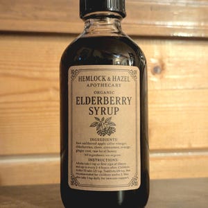 Pu&ograve; includere: Una bottiglia di vetro trasparente di sciroppo di sambuco biologico Hemlock & Hazel Apothecary. L'etichetta elenca ingredienti come bacche di sambuco e miele. La bottiglia ha un tappo nero ed &egrave; piena di un liquido scuro.