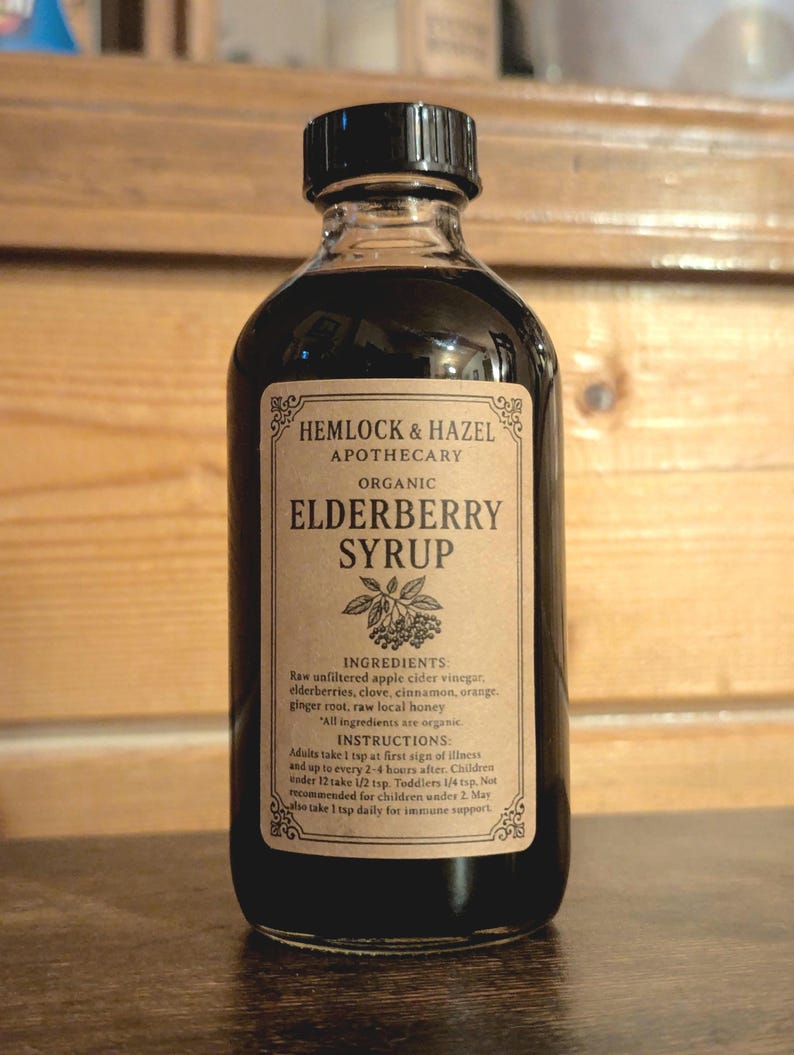Pu&ograve; includere: Una bottiglia di vetro trasparente di sciroppo di sambuco biologico Hemlock & Hazel Apothecary. L'etichetta elenca ingredienti come bacche di sambuco e miele. La bottiglia ha un tappo nero ed &egrave; piena di un liquido scuro.