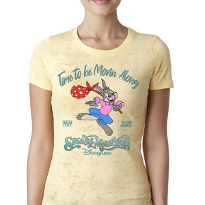 Peut inclure: Un t-shirt jaune délavé avec un lapin de dessin animé tenant un bâton et un sac à pois rouges. Le texte "Time to be Movin' Along" est écrit en bleu au-dessus du lapin. Le texte "1989" et "2023" est écrit en bleu de chaque côté du lapin. Le texte "Splash Mountain Disneyland" est écrit en bleu sous le lapin.