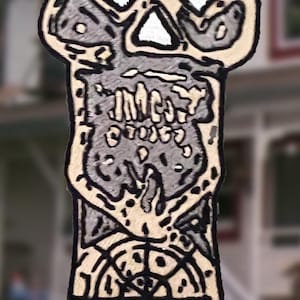 May include: A vertical embroidered patch featuring a stylized skull at the top with dark gray and cream-colored thread. Below the skull, the word "INTO" is stitched in black. The bottom section displays a circular geometric compass-like design.