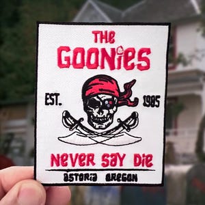 May include: A rectangular embroidered patch with a white background and black border. It features a red skull wearing a bandana, crossed swords, and the text: THE GOONIES, EST. 1985, NEVER SAY DIE, and ASTORIA OREGON in red and black lettering.