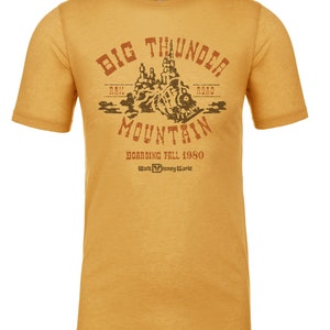 Puede incluir: Una camiseta amarilla con un gr&aacute;fico de estilo vintage de Big Thunder Mountain Railroad. El gr&aacute;fico presenta una monta&ntilde;a marr&oacute;n con una v&iacute;a de tren y el texto "Big Thunder Mountain" en un arco marr&oacute;n. El texto "Rail Road" est&aacute; a ambos lados de la monta&ntilde;a. El texto "Boarding Fall 1980" y "Walt Disney World" est&aacute; debajo de la monta&ntilde;a.