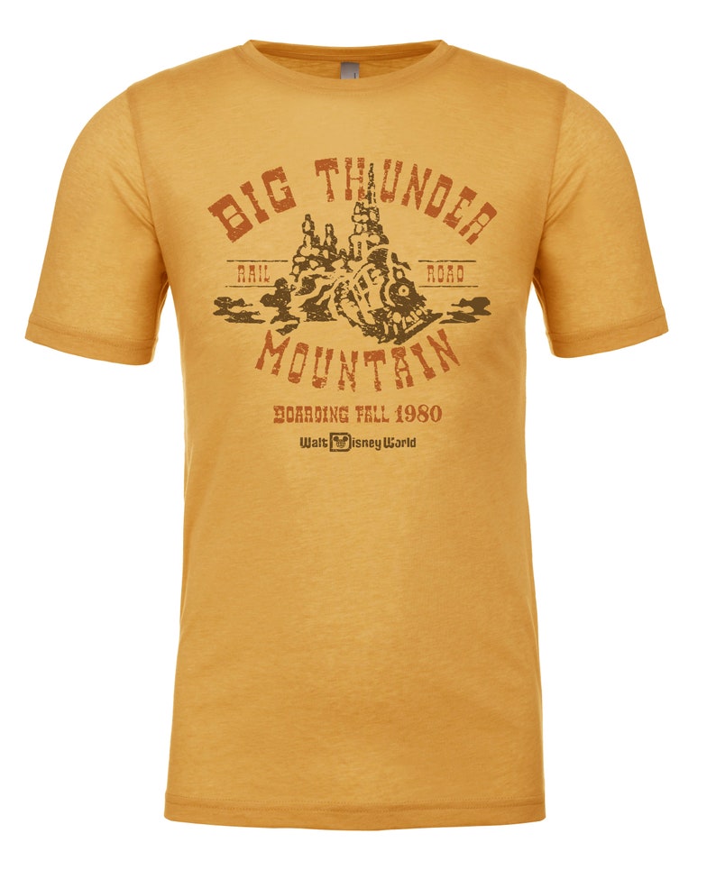 Puede incluir: Una camiseta amarilla con un gr&aacute;fico de estilo vintage de Big Thunder Mountain Railroad. El gr&aacute;fico presenta una monta&ntilde;a marr&oacute;n con una v&iacute;a de tren y el texto "Big Thunder Mountain" en un arco marr&oacute;n. El texto "Rail Road" est&aacute; a ambos lados de la monta&ntilde;a. El texto "Boarding Fall 1980" y "Walt Disney World" est&aacute; debajo de la monta&ntilde;a.