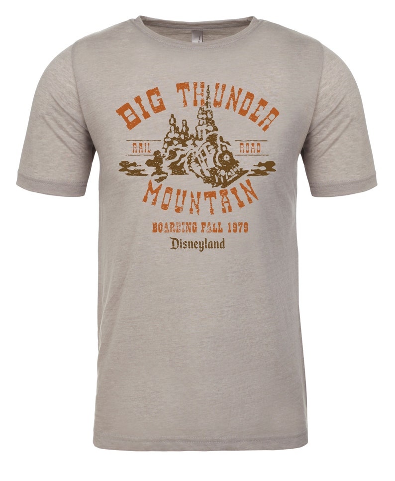 Puede incluir: Una camiseta gris jaspeada con un gr&aacute;fico de estilo vintage de Big Thunder Mountain Railroad. El gr&aacute;fico presenta una silueta marr&oacute;n del paseo con el texto "Big Thunder Mountain" en letras marrones y naranjas. El texto "Rail Road" est&aacute; en letras marrones m&aacute;s peque&ntilde;as por encima del paseo, y el texto "Boarding Fall 1979 Disneyland" est&aacute; en letras marrones m&aacute;s peque&ntilde;as por debajo del paseo.
