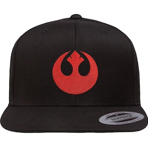 Op de afbeelding: Zwarte snapback pet met een rood geborduurd Rebel Alliance symbool. De pet heeft een platte klep en een druksluiting aan de achterkant. De merknaam "Snapback" is zichtbaar.