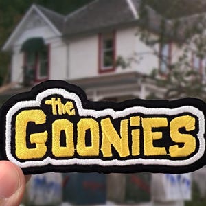 May include: An embroidered patch featuring the text "the GOONIES" in yellow thread with a white outline on a black background. The patch has a thick black border and a textured surface created by the embroidery stitching process.