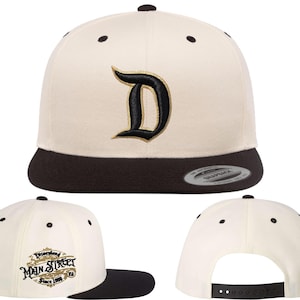 Puede incluir: Gorra de béisbol color crema con visera negra y una letra "D" bordada en negro con contorno dorado. La gorra también presenta el texto "Disneyland Main Street Since 1955" bordado en el lateral.