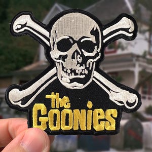 May include: An embroidered patch featuring a white skull and crossbones on a black background. Below the skull, the text "The GOONIES" is stitched in bold yellow lettering. The patch has a black border and a distinct, textured thread finish.