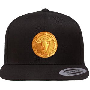 Peut inclure: Casquette noire avec un écusson doré brodé. L'écusson est circulaire avec un motif d'éclair stylisé. La casquette a une visière plate et une fermeture à pression. La marque "The Classics" est visible sur la visière.