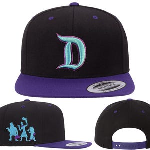 May include: Black snapback cap with a purple peak and a teal and purple embroidered "D" on the front. The side features a teal cartoon graphic. The cap has a snap closure for adjustable sizing.