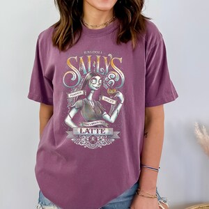 Puede incluir: Una camiseta morada con un gr&aacute;fico de estilo vintage de Sally de Pesadilla Antes de Navidad. El gr&aacute;fico muestra a Sally sosteniendo un latte de calabaza especiado y el texto "Ragdoll Sally's Pumpkin Spiced Latte".