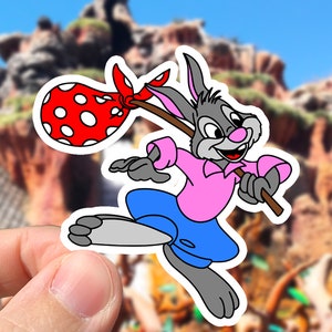 May include: A cartoon rabbit wearing a pink shirt and blue shorts is holding a stick with a red and white polka dot bag attached to it. The rabbit is running and has a determined expression on its face.