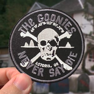 May include: A circular black embroidered patch featuring a white skull and crossbones design. The text reads THE GOONIES at the top and NEVER SAY DIE at the bottom, with the coordinates 46 11' 42"N 123 50' 72"W and ASTORIA, OR printed in white.