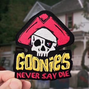 May include: A black embroidered patch featuring a white skull wearing a red pirate hat with an eye patch. Below the skull, the word GOONIES is stitched in yellow, followed by the text NEVER SAY DIE in red, all set against a black background.