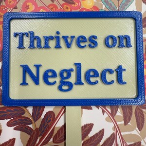 May include: A blue and beige garden sign with the text "Thrives on Neglect" in blue lettering.